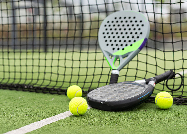 Zwei Padelschläger einer steht an einem Netz, einer liegt davor. Herum liegen drei Tennisbälle