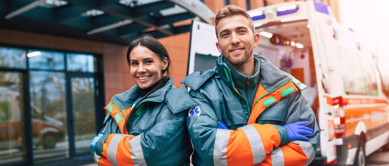 Frau und Mann in orange-blauer Kleidung haben beide die Arme verschränkt. Im Hintergrund ist ein Krankenwagen zu sehen