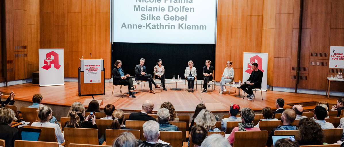 Ein holzvertäfelter Saal, auf dem Podium sitzen sieben Personen. Aufsteller mit dem Apotheken-A, rotes Flatterband.