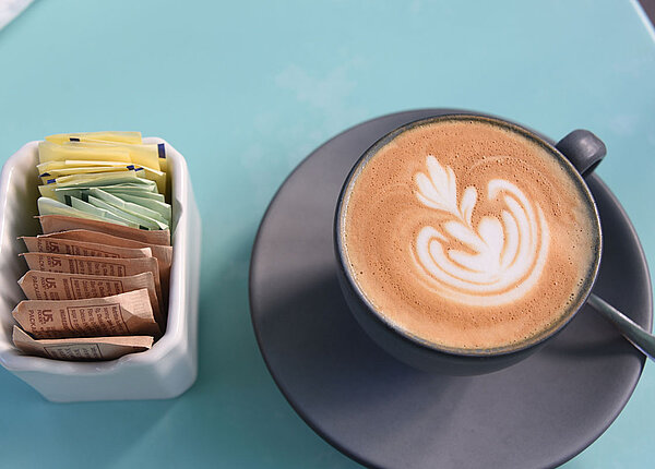 Eine Tasse Kaffe mit Milchschaum. Links daneben ein Gefäß mit Süßungsmitteln in Sachets in drei verschiedenen Farben.