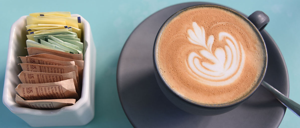 Eine Tasse Kaffe mit Milchschaum. Links daneben ein Gefäß mit Süßungsmitteln in Sachets in drei verschiedenen Farben.