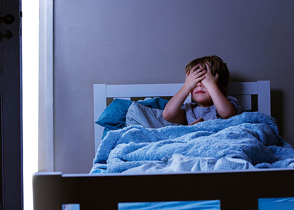 Ein Junge von etwa vier Jahren sitzt im Dunklen in seinem Bett und hält sich die Augen zu.