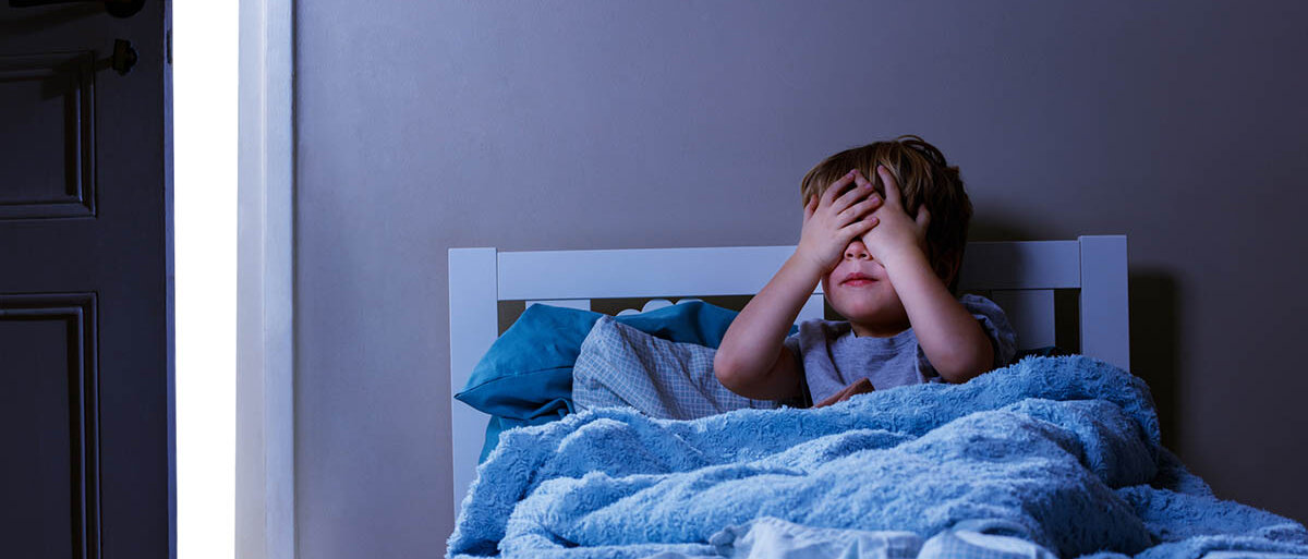 Ein Junge von etwa vier Jahren sitzt im Dunklen in seinem Bett und hält sich die Augen zu.