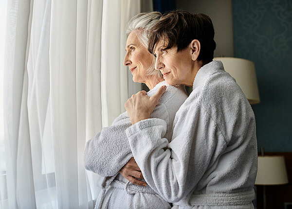 Wundheilung beschleunigen mit dem Kuschelhormon Oxytocin Zwei Seniorinnen im Bademantel stehen am Fenster und kuscheln.