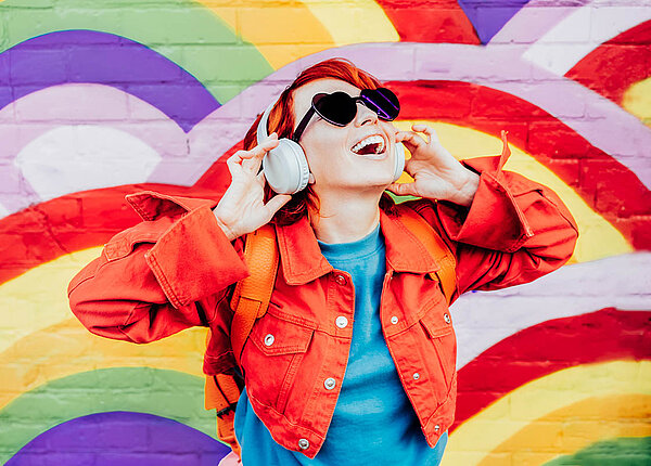 Synästhesie-Wahrnehmungen betreffen alle Sinne. Es gibt verschiedene Formen, zum Beispiel die Graphem-Farb-Anästhesie. Eine junge Frau hört Musik über ihre Kopfhörer. Sie ist bunt gekleidet, hat knallrote Haare und die Mauer hinter ihr ist mit Regenbögen bemalt.