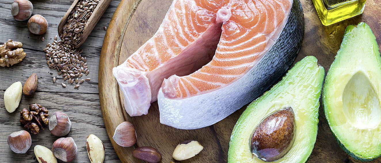 Auf einem runden Holzteller liegt eine gut fingerdicke Scheibe roher Lachs und eine aufgeschnittene Avocado, außerdem eine kleine Phiole voll Öl. Neben dem Teller liegen verschiedene Nüsse, Kerne und Samen.