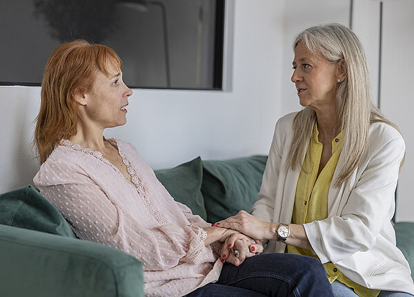 Wann und wem man von einer Krebsdiagnose erzählen sollte Zwei Frauen sitzen gemeinsam auf einer Couch, haben die Hände übereinandergelegt, sitzen sich gegenüber, sprechen offen miteinander, ernste und einfühlsame Stimmung.