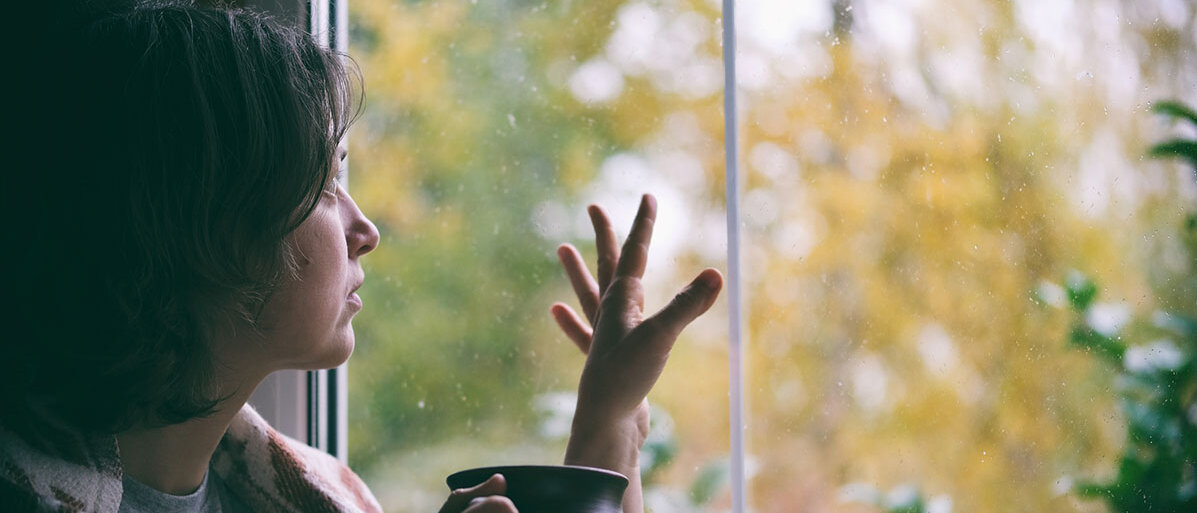 Frau sitzt am Fenster mit einer Tasse in der Hand und schaut raus auf die herbstliche Landschaft und Regentropfen sind an der Fensterscheibe zu sehen.