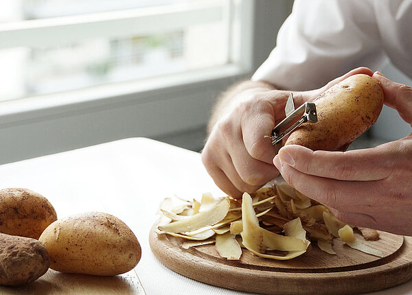 Mann schält mit einem Schälmesser eine Kartoffel auf einem Brett. Davor liegen drei weitere Kartoffeln.