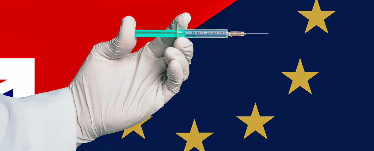 Eine Hand mit weißen Einmalhandschuhen hält eine Spritze mit der Aufschrift "Covid-19 Vaccine", im Hintergrund sind die Flaggen von Großbritannien und der Europäischen Union.