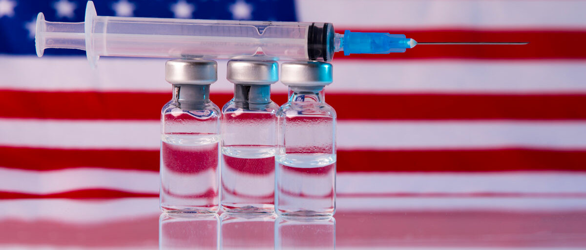 Drei Impfstofffläschchen und eine Spritze vor der US-amerikanischen Flagge