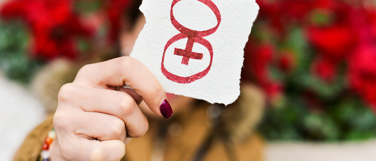 eine Hand mit lackierten Nägeln hält einen Zettel hoch, darauf: eine Acht mit integriertem Frauenzeichen