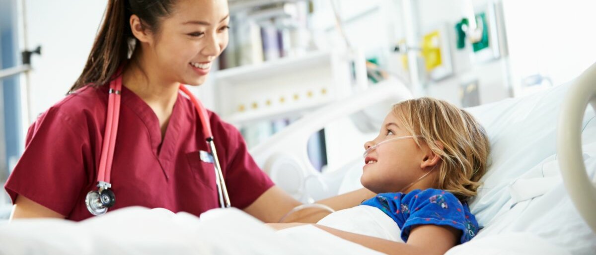 Ein Kind liegt in einem Krankenbett, daneben steht eine lächelnde Krankenschwester.