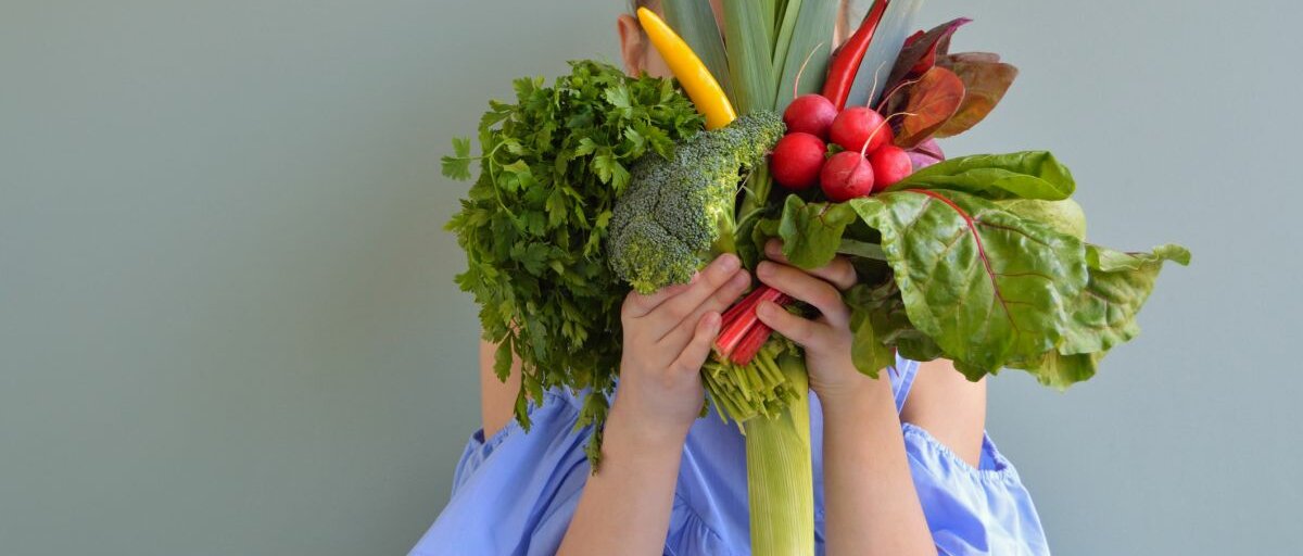 Eine junge Frau, die verschiedenes Gemüse vor ihr Gesicht hält.