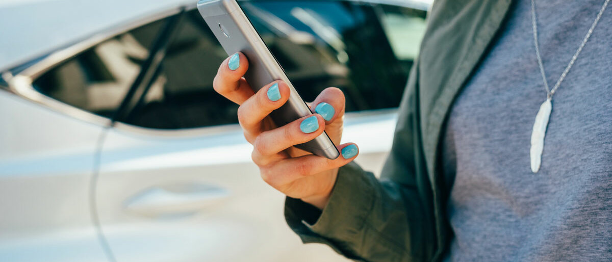 Eine Frau steht vor einem weißen Auto und tippt auf ihrem Smartphone