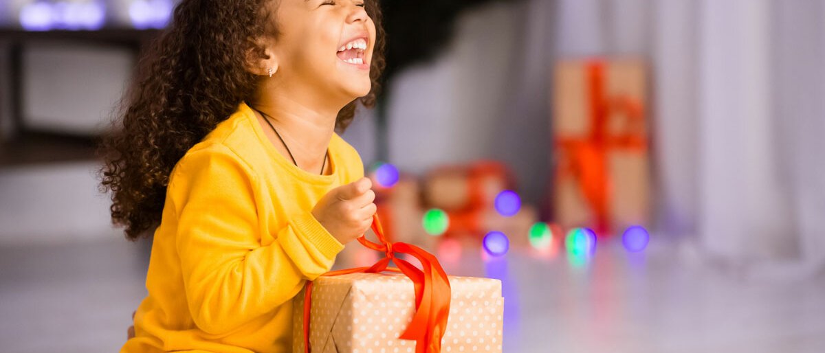 Ein kleines Mädchen sitzt vor dem Weihnachtsbaum, packt ein Geschenk aus und lächelt strahlend.