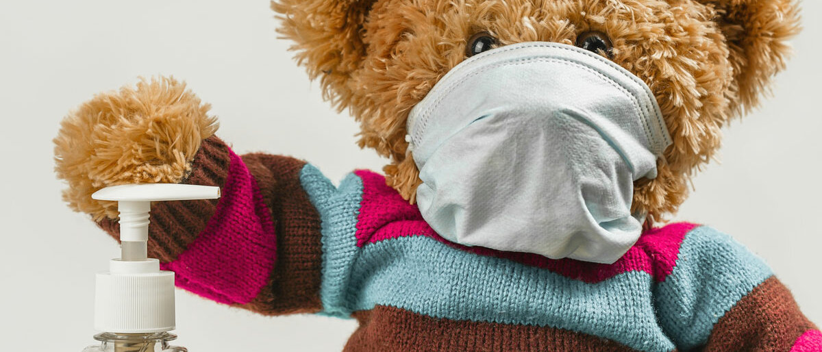 Ein Teddy trägt MNS und hat die Pfote auf einem Desinfektionsmittelspender liegen.