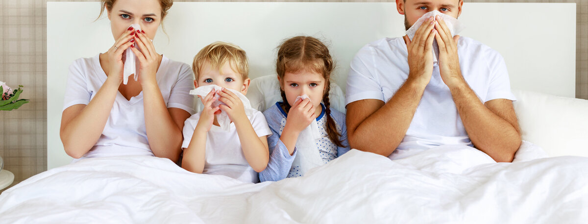 Mutter, Vater und zwei Kinder sitzen krank im Familienbett.