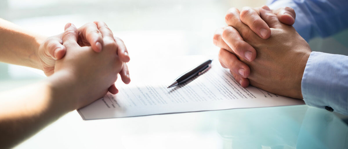 Zwei Paar Hände sind über einem Vertrag gefaltet. Ein Stift liegt zum Unterschreiben bereit.