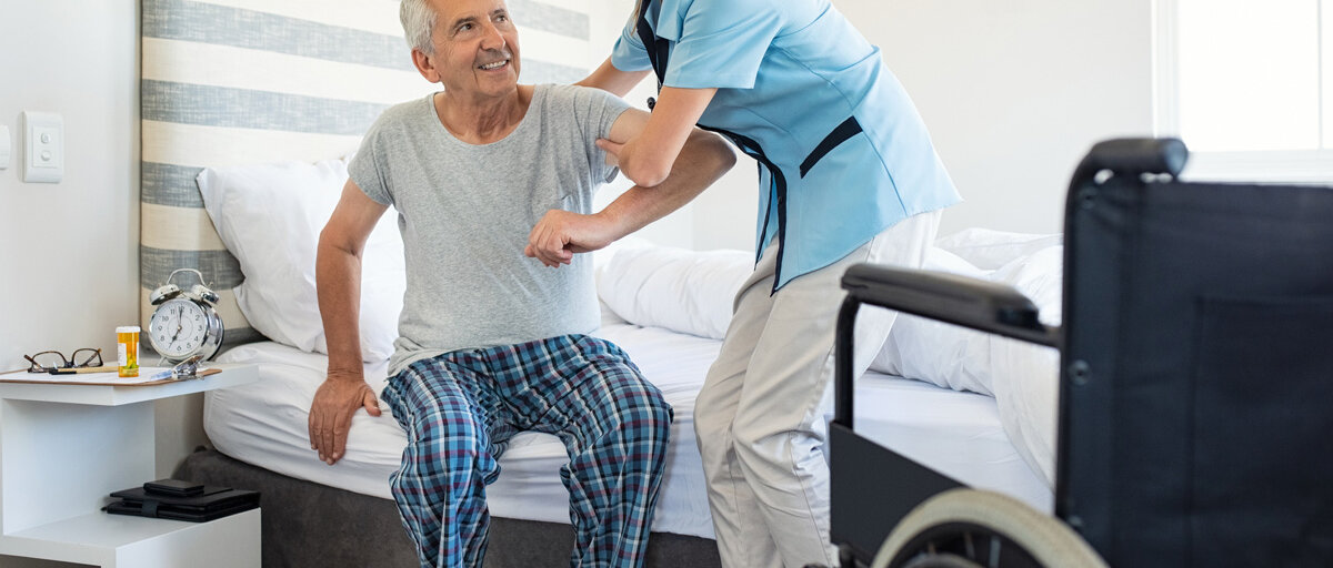 Eine Pflegerin hilft einem älteren Mann aus seinem Bett in den Rollstuhl