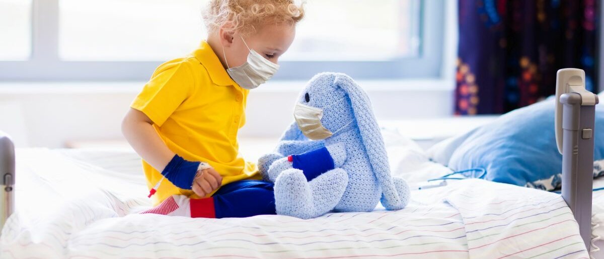 Ein Kind sitzt mit seinem Stoffhasen im Bett eines Krankenhauses und trägt einen Mundschutz.