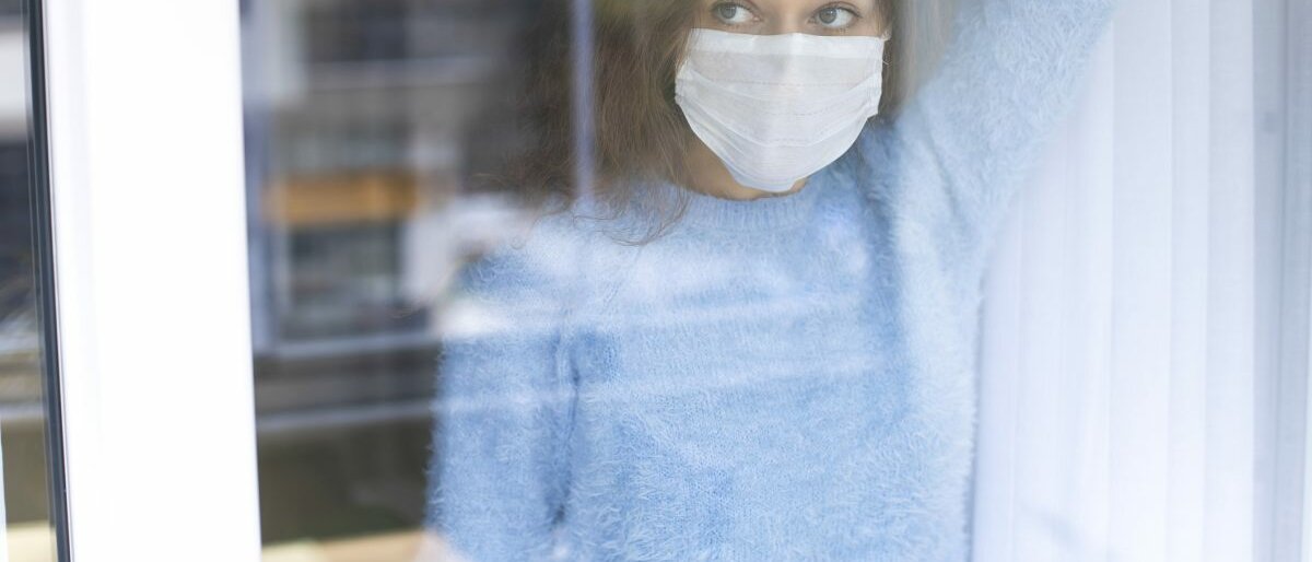 unge Frau in Quarantäne trägt eine Maske und schaut durch das Fenster.